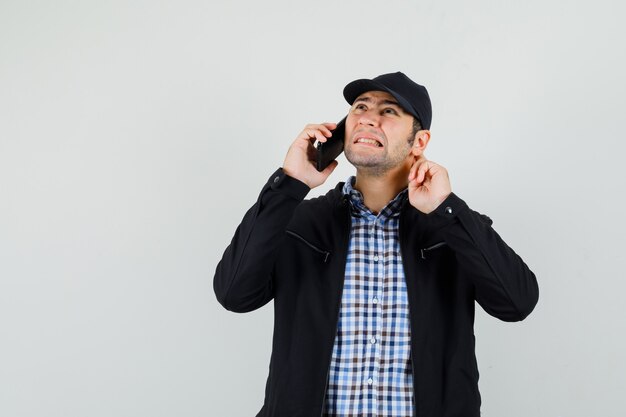 Hombre joven en camisa, chaqueta, gorra oyendo mal mientras habla por teléfono móvil