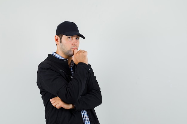 Hombre joven en camisa, chaqueta, gorra de pie en pose de pensamiento y mirando serio