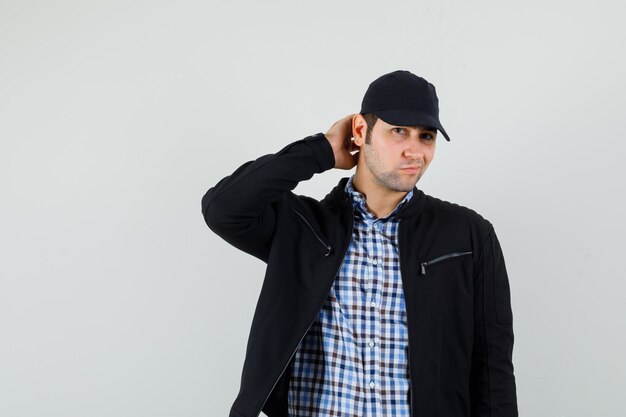 Hombre joven en camisa, chaqueta, gorra posando mientras se rasca la cabeza y se ve elegante