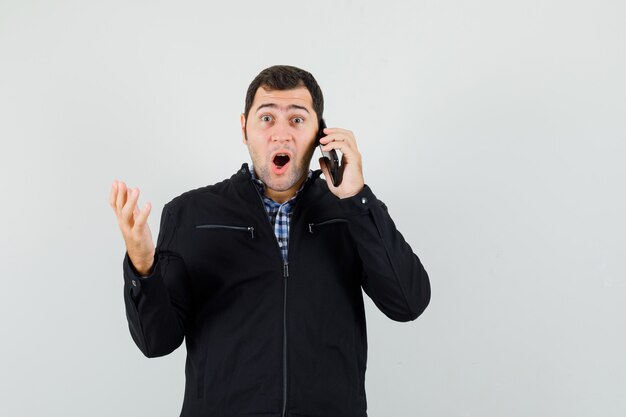 Hombre joven en camisa, chaqueta hablando por teléfono móvil y mirando asombrado