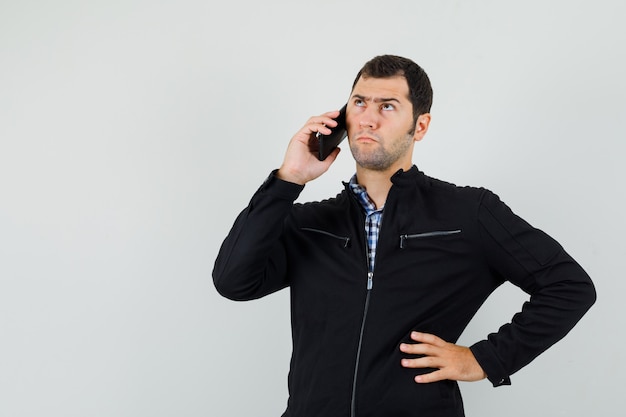 Hombre joven en camisa, chaqueta hablando por teléfono móvil y mirando indeciso