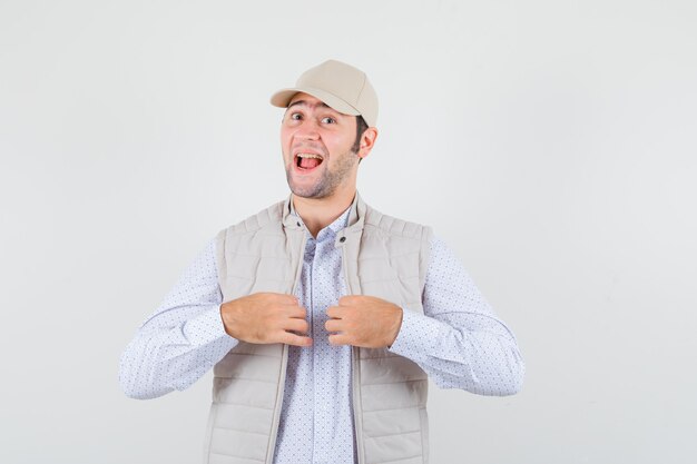 Hombre joven en camisa, chaqueta sin mangas, gorra cogidos de la mano en el cuello y mirando satisfecho, vista frontal.