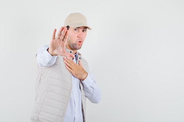 Hombre joven en camisa, chaqueta sin mangas, gorra levantando la mano mientras rechaza algo y parece reacio, vista frontal.