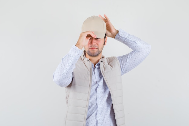 Hombre joven en camisa, chaqueta sin mangas con gorra y mirando confiado, vista frontal.