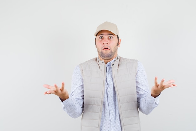 Hombre joven en camisa, chaqueta sin mangas, gorra con las palmas abiertas a un lado con actitud interrogativa y aspecto preocupado, vista frontal.