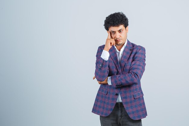 Hombre joven en camisa, chaqueta, pantalones de pie en pose de pensamiento y mirando sensible, vista frontal.