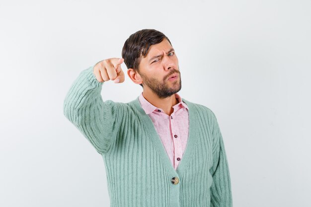 Hombre joven en camisa, chaqueta de punto apuntando hacia adelante y mirando confiado, vista frontal.