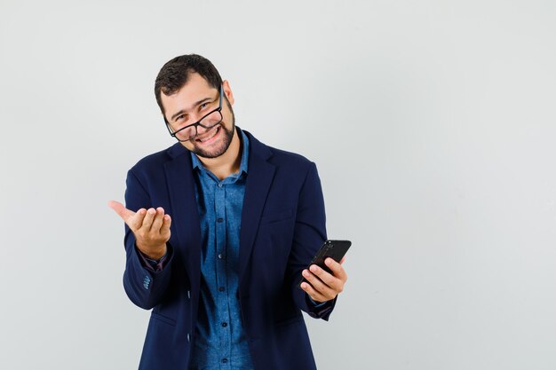 Hombre joven en camisa, chaqueta sosteniendo teléfono móvil y sonriendo, vista frontal.