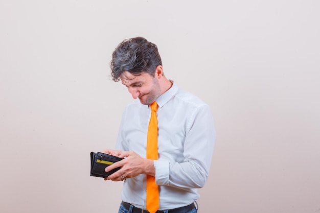 Hombre joven en camisa, corbata, jeans mirando a la billetera y mirando alegre