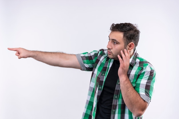 Hombre joven en camisa a cuadros apuntando con el dedo hacia el lado mirando confundido mientras habla por teléfono móvil de pie sobre la pared blanca