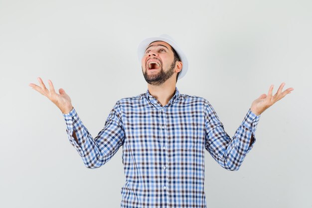 Hombre joven con camisa a cuadros, sombrero extendiendo las manos mientras mira hacia arriba y se ve feliz, vista frontal.