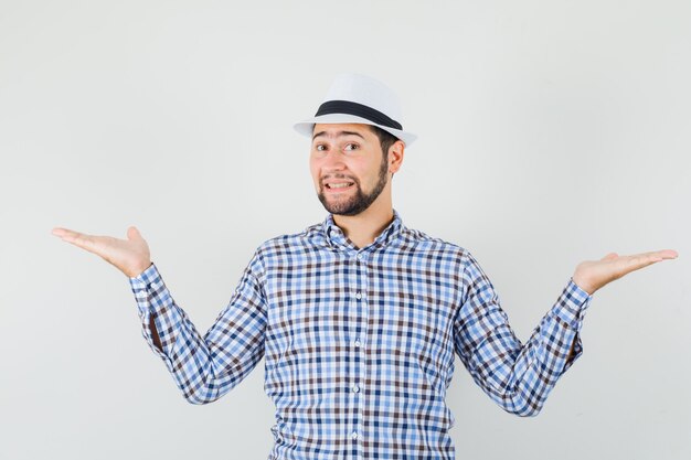 Hombre joven en camisa a cuadros, sombrero haciendo gesto de escalas y mirando alegre, vista frontal.