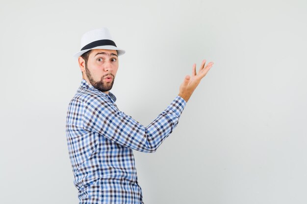 Hombre joven con camisa a cuadros, sombrero levantando la mano y mirando sorprendido.