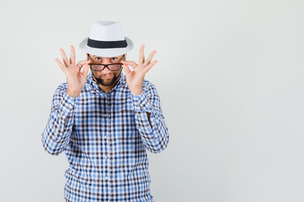 Hombre joven con camisa a cuadros, sombrero mirando atentamente por encima de las gafas y mirando enfocado, vista frontal.