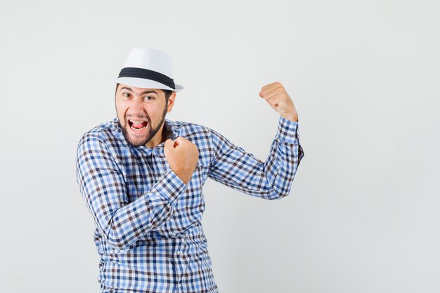 Hombre joven con camisa a cuadros, sombrero mostrando gesto de ganador y mirando feliz, vista frontal.