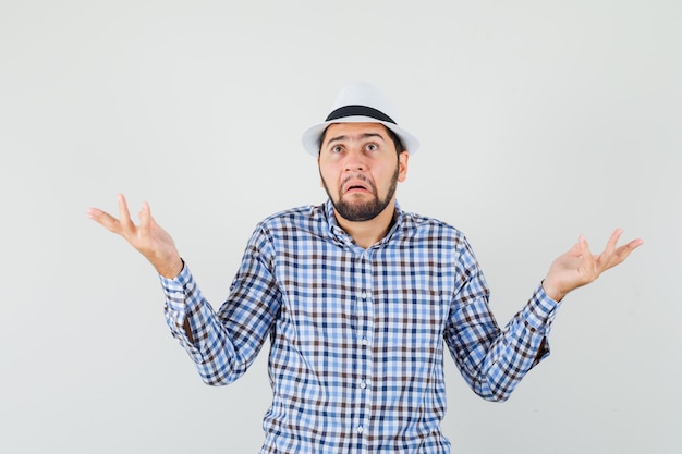 Hombre joven en camisa a cuadros, sombrero mostrando gesto de impotencia y mirando confundido, vista frontal.