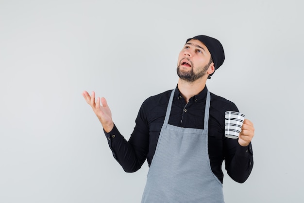 Hombre joven en camisa, delantal sosteniendo una taza de bebida y mirando confundido, vista frontal.