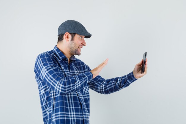 Hombre joven en camisa, gorra agitando la mano en el chat de video y mirando alegre, vista frontal.