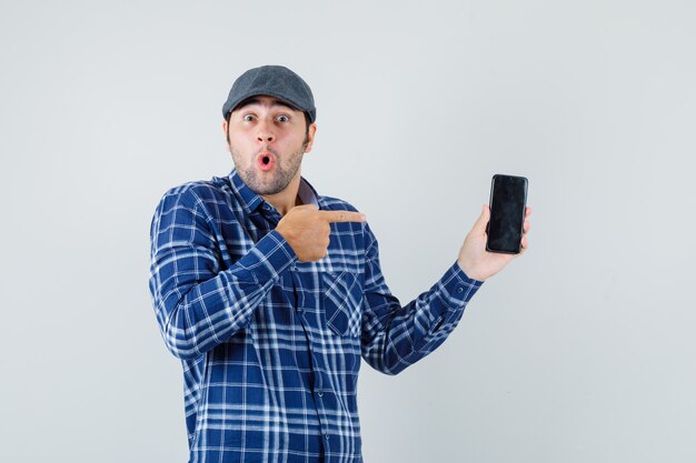 Hombre joven en camisa, gorra apuntando al teléfono móvil y mirando sorprendido, vista frontal.