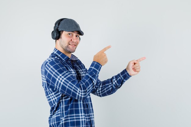 Hombre joven en camisa, gorra apuntando a la esquina superior derecha y mirando alegre, vista frontal.