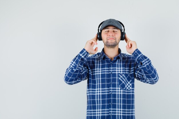 Hombre joven en camisa, gorra disfrutando de la música con auriculares y mirando encantado, vista frontal.