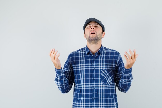 Hombre joven en camisa, gorra levantando las manos en gesto de oración y mirando esperanzado, vista frontal.