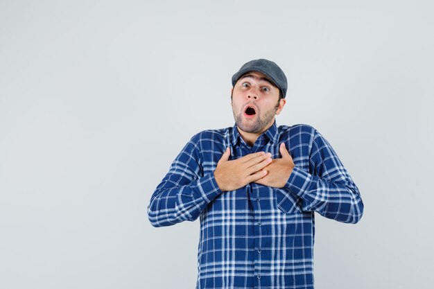 Hombre joven en camisa, gorra de la mano en el pecho y mirando asustado, vista frontal.