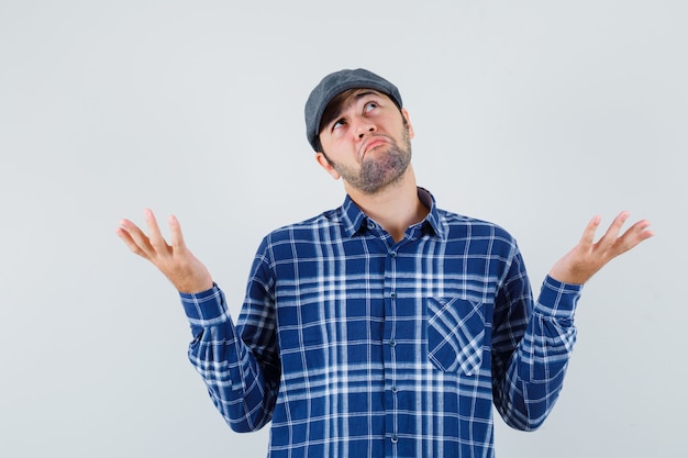 Foto gratuita hombre joven en camisa, gorra mostrando gesto de impotencia y mirando confundido, vista frontal.