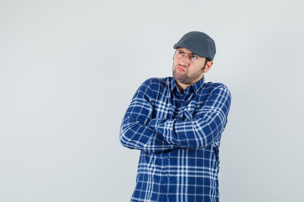 Hombre joven en camisa, gorra de pie con los brazos cruzados y mirando vacilante, vista frontal.