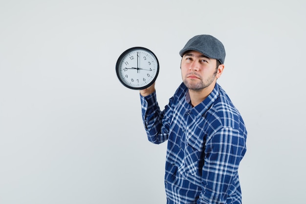 Hombre joven en camisa, gorra sosteniendo el reloj de pared y mirando pensativo.
