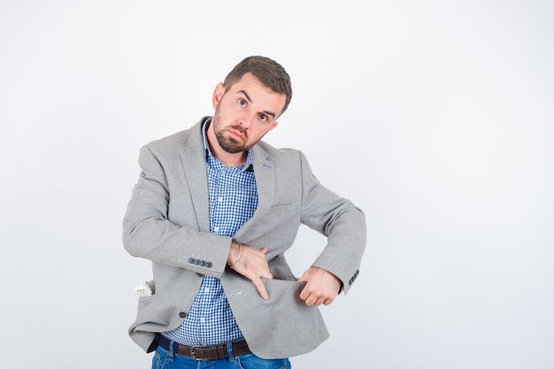 Hombre joven en camisa, jeans, chaqueta de traje abriendo el bolsillo de la chaqueta con las manos y mirando serio, vista frontal.