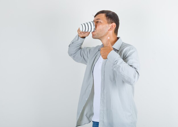 Hombre joven en camisa, jeans tomando café con el pulgar hacia arriba