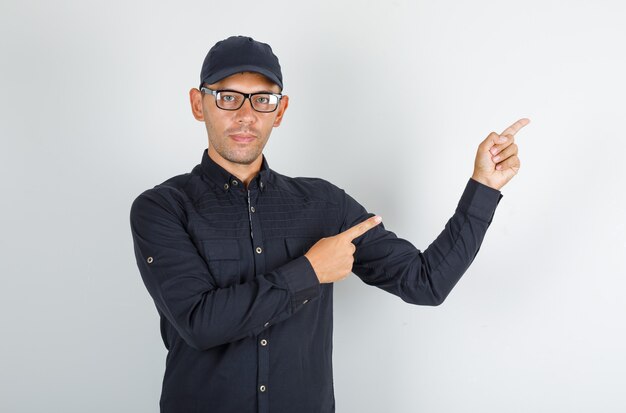 Hombre joven en camisa negra con gorra, gafas apuntando con el dedo lejos