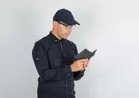 Foto gratuita hombre joven en camisa negra con gorra, gafas mirando a través del cuaderno