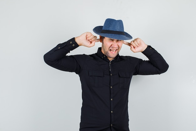 Hombre joven con camisa negra, sombrero tapando las orejas con los dedos y mirando molesto