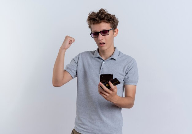 Hombre joven en camisa de polo gris mirando la pantalla de su teléfono inteligente con tarjeta de crédito apretando el puño feliz y emocionado de pie sobre la pared blanca