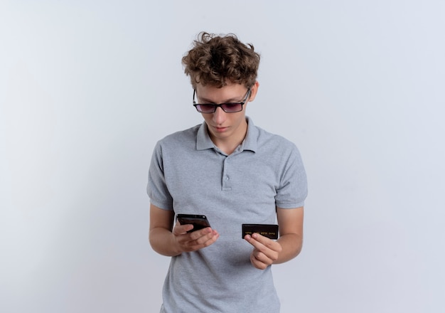 Hombre joven en camisa polo gris mirando la pantalla de su teléfono inteligente con tarjeta de crédito confundido de pie sobre la pared blanca