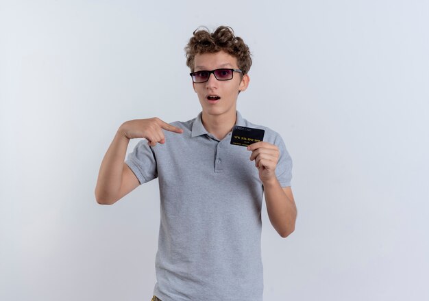 Hombre joven en camisa polo gris que muestra la tarjeta de crédito apuntando con el dedo a que se sorprende de pie sobre la pared blanca