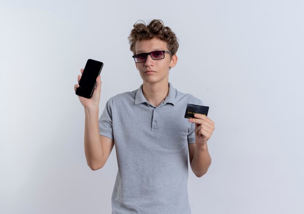 Hombre joven en camisa polo gris que muestra el teléfono inteligente con tarjeta de crédito con cara seria de pie sobre la pared blanca