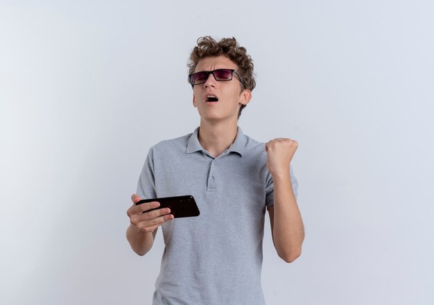 Hombre joven en camisa polo gris con smartphone apretando el puño feliz y emocionado de pie sobre la pared blanca