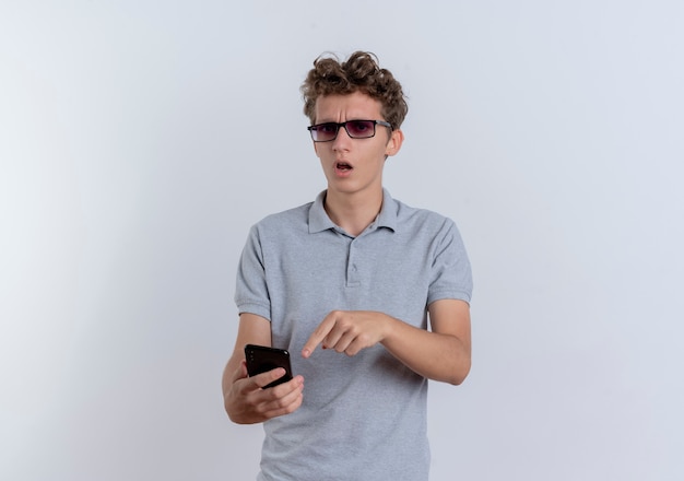 Hombre joven en camisa polo gris con smartphone apuntando con el dedo índice a confundirse de pie sobre la pared blanca