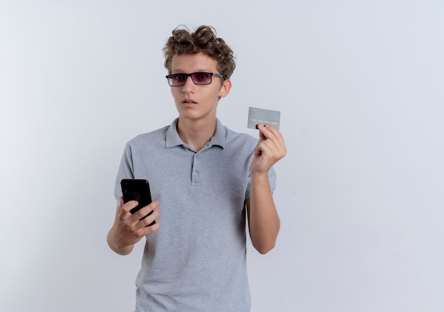Hombre joven en camisa polo gris con smartphone mostrando tarjeta de crédito confundido parado sobre pared blanca