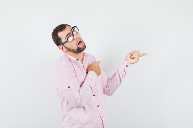 Hombre joven en camisa rosa apuntando hacia afuera y mirando divertido