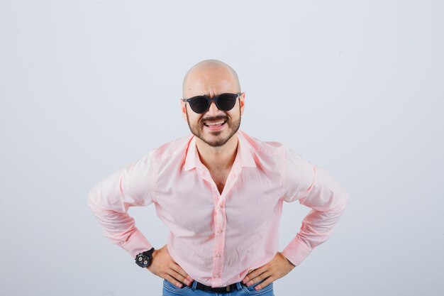 Hombre joven con camisa rosa, jeans, gafas de sol pidiendo algo con las manos en la cintura y mirando curioso, vista frontal.