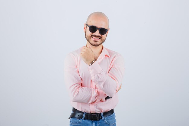 Hombre joven en camisa rosa, jeans, gafas de sol sonriendo y mirando genial, vista frontal.