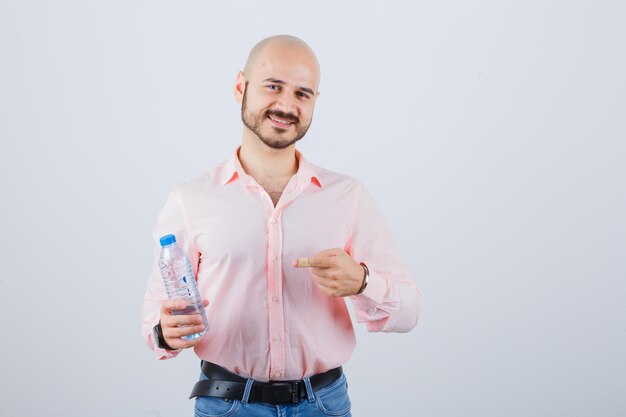 Hombre joven en camisa rosa, jeans sosteniendo una botella de agua mientras apunta hacia ella, vista frontal.