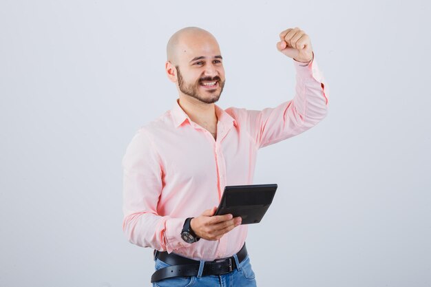 Hombre joven en camisa rosa, jeans sosteniendo la calculadora mientras muestra un gesto de éxito y luce alegre, vista frontal.