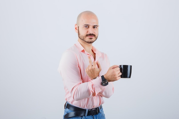 Hombre joven en camisa rosa, jeans sosteniendo la taza mientras muestra el dedo medio, vista frontal.