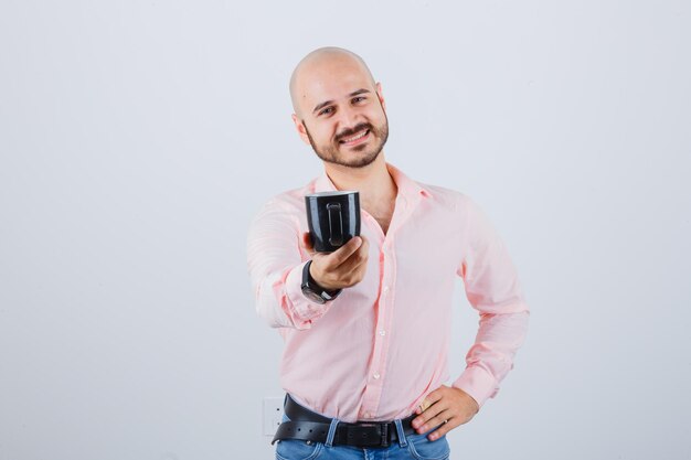 Hombre joven en camisa rosa, jeans sosteniendo la taza mientras sonríe, vista frontal.