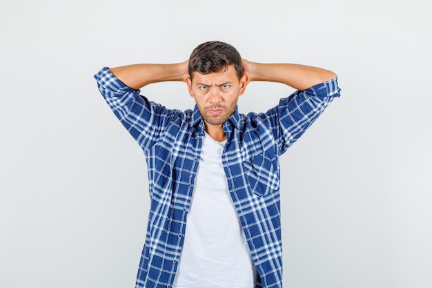 Foto gratuita hombre joven en camisa tomados de la mano detrás de la cabeza y mirando nervioso, vista frontal.